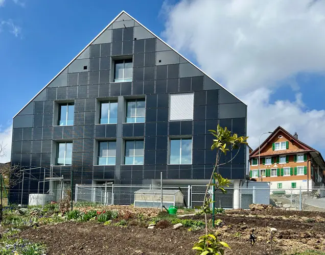Modern house with PV panels on the facade, a freshly planted garden, a rustic house in the background, and a few clouds in the sky.