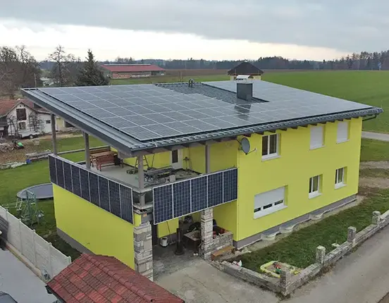 Yellow house with solar panels on roof and balcony, set in rural landscape with fields and cloudy sky