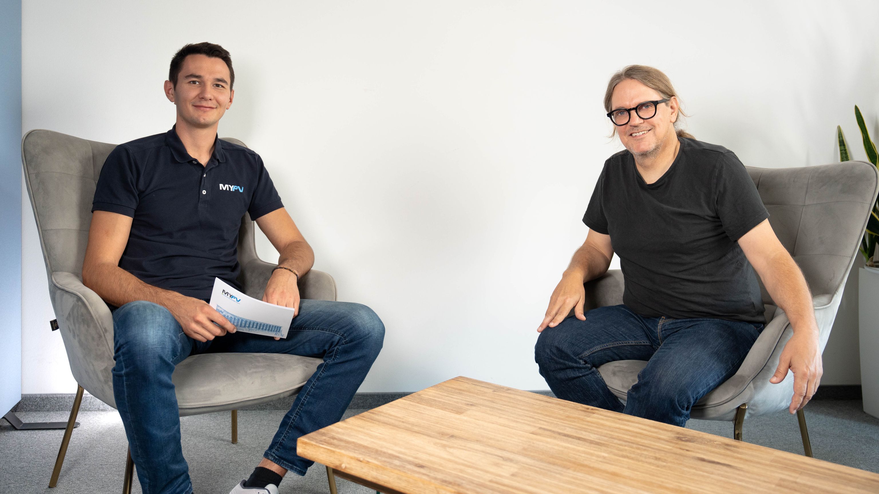 Two men sitting in armchairs, smiling. One holds papers, the other has long hair. A wooden table is between them, and a plant is in the background.