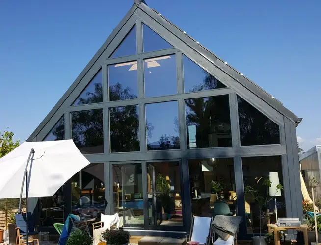 Modern A-frame house with large glass windows, outdoor furniture, and white umbrella under clear blue sky.
