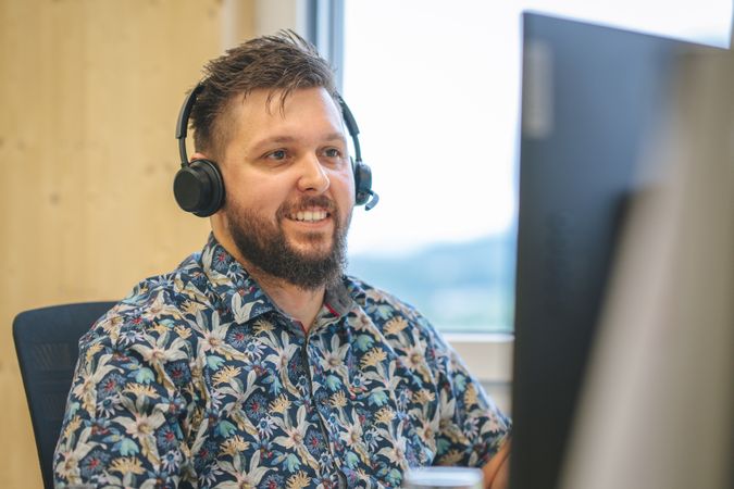 Man with a beard wearing headphones and a floral shirt, smiling while looking at a computer screen in a bright office.