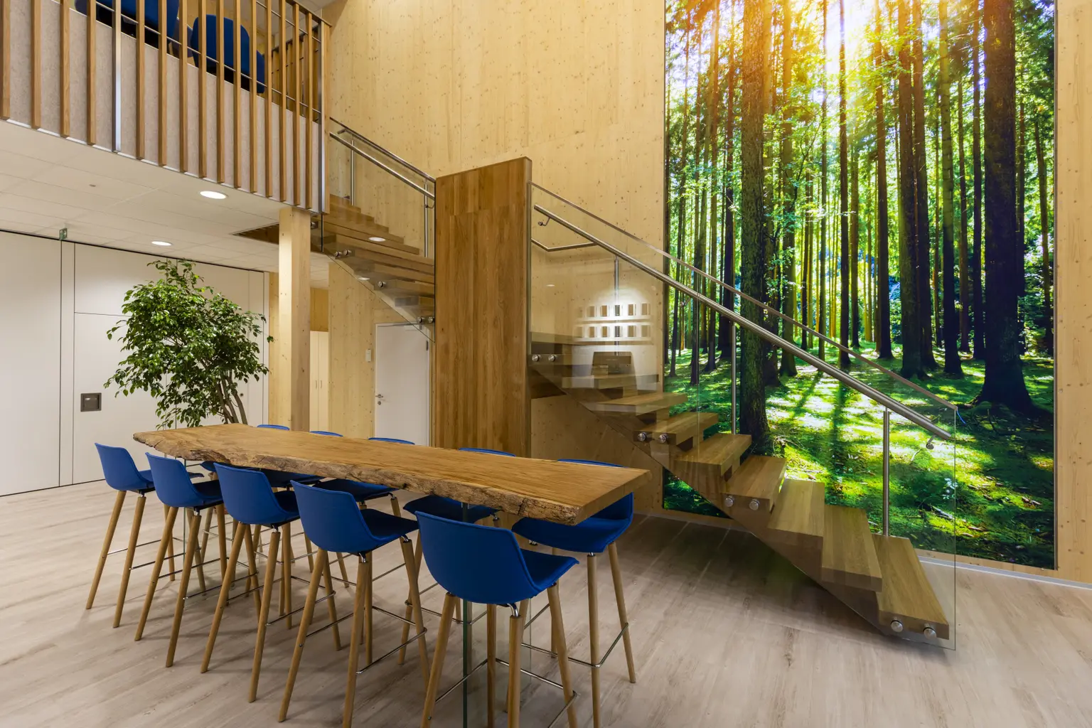 Foyer with stairs view. In front, a standing table with blue chairs. A large forest picture hangs on the wall by the stairs.