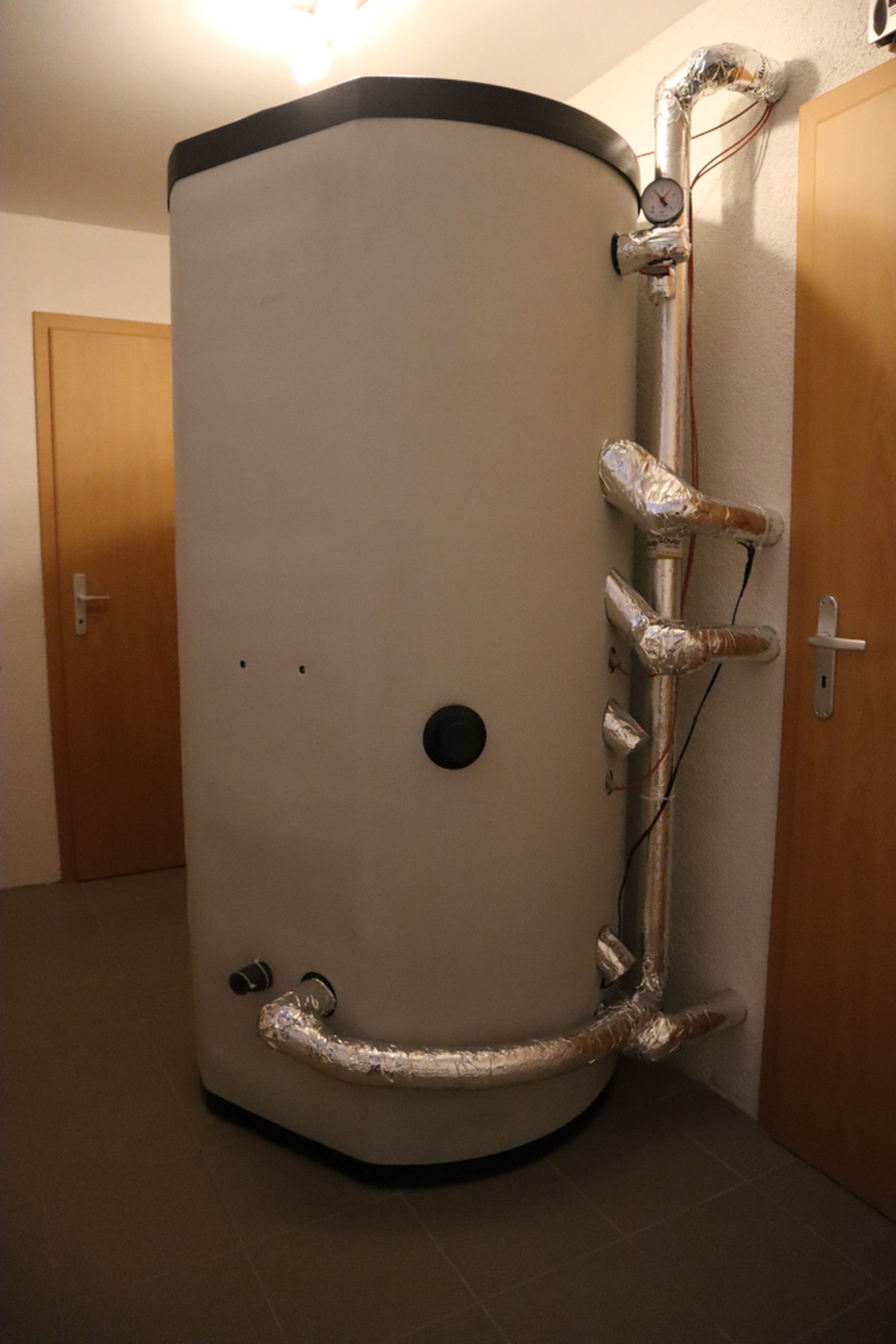A large, cylindrical water heater tank with insulated pipes attached stands in a room between two wooden doors.