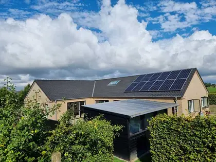 House with solar panels on the roof, a garden shed in front, surrounded by lush shrubs
