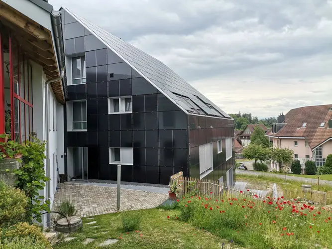 Modern house with dark facade next to another house, garden featuring vibrant red poppies and greenery.