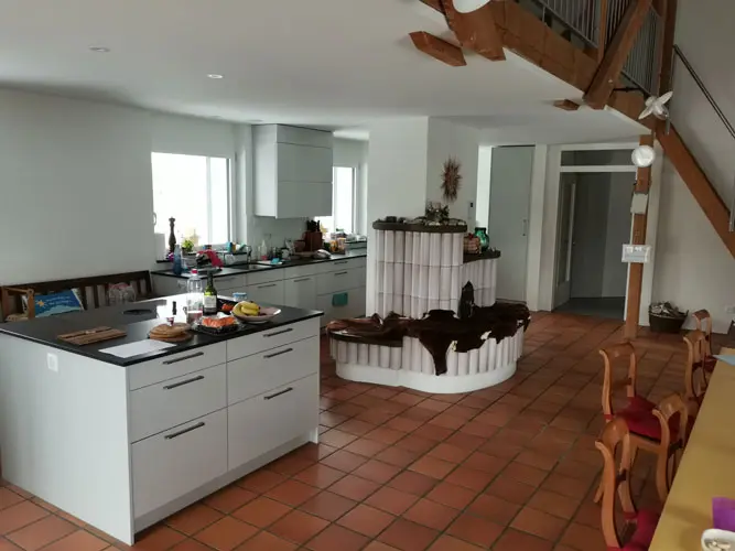 Modern kitchen with white cabinets, dark countertop, rustic tiled stove in the center, and reddish-brown tiled floor.