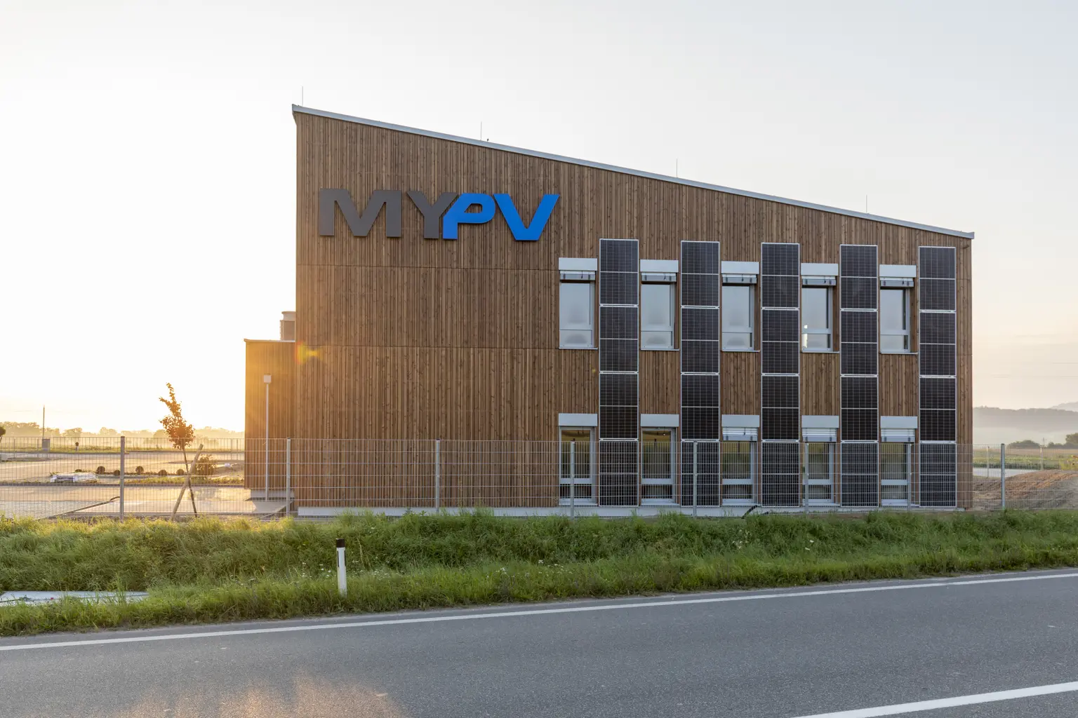 The company building is shown from the side with logo and PV panels. The sun shines behind, street, grass, and parking in front.