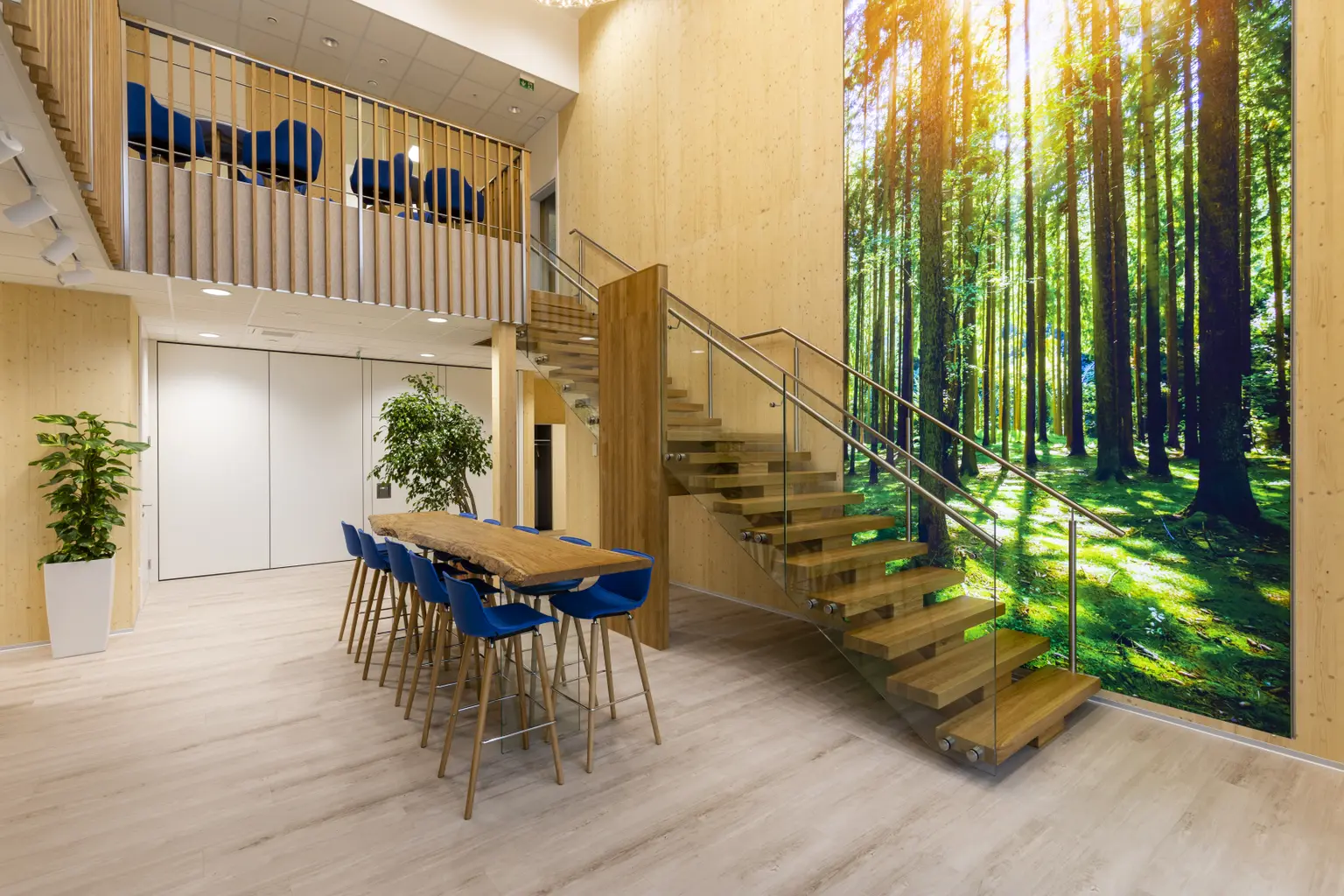 Foyer with stairs view. In front, a standing table with blue chairs. A large forest picture hangs on the wall by the stairs.