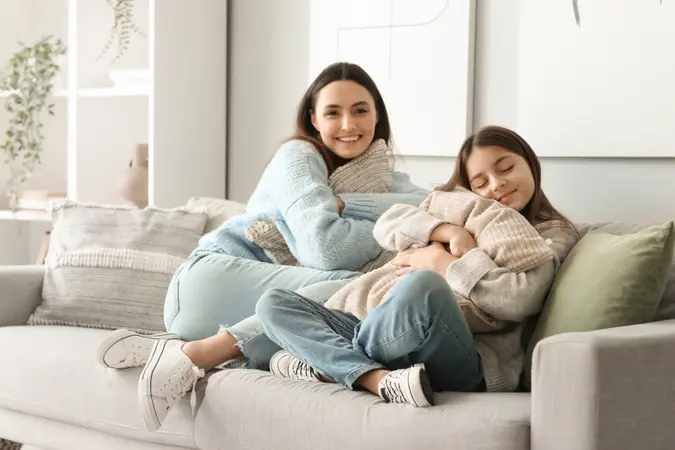 Two women in cozy sweaters sitting on a sofa, one smiling at the camera and the other resting with eyes closed while hugging a pillow.