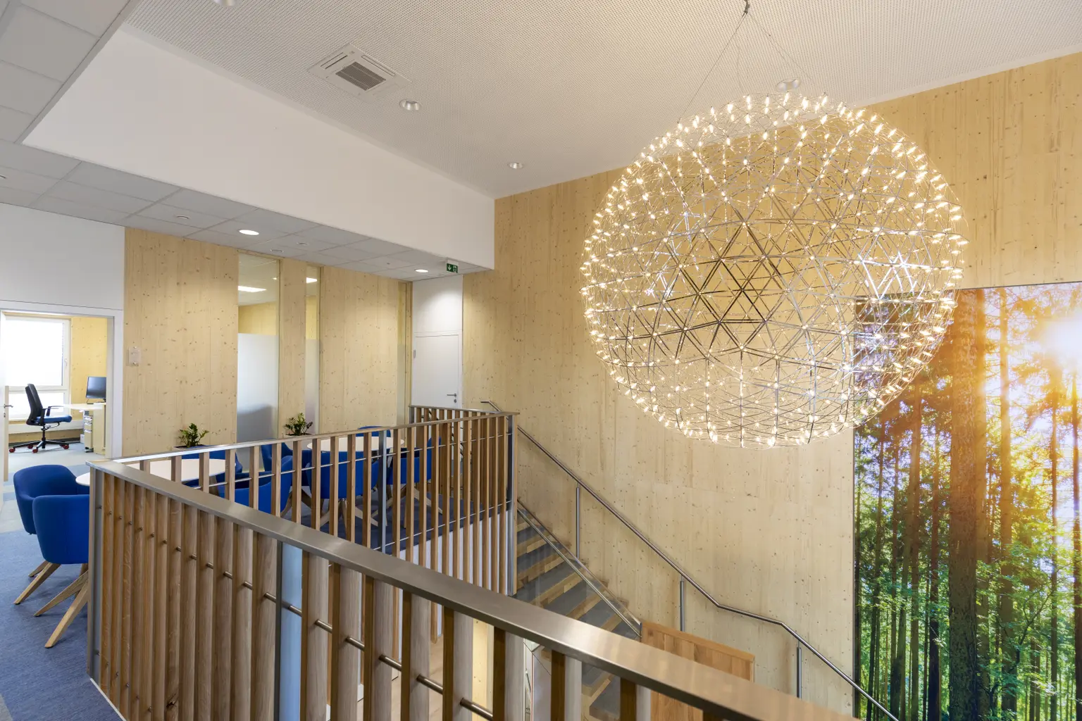 Foyer viewed from above with wooden walls and large sun-like lampshade. Seating on left, forest picture by stairs on right.