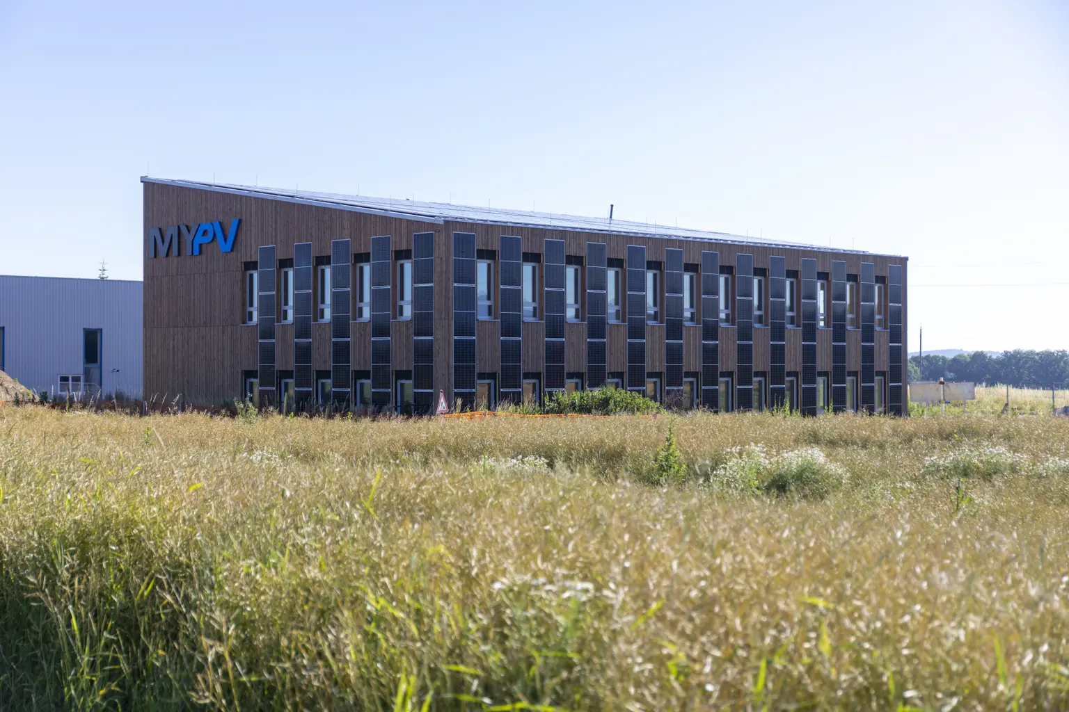 The company building is seen from afar, with PV panels on the roof and facade. A barley field stretches out in the foreground.
