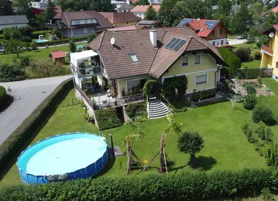 Aerial view of a suburban house with brown roof, green lawn, circular above-ground pool, and neighboring houses