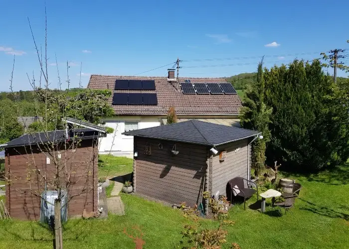 House with solar panels on the roof, surrounded by grass, trees, and two wooden sheds under clear blue sky