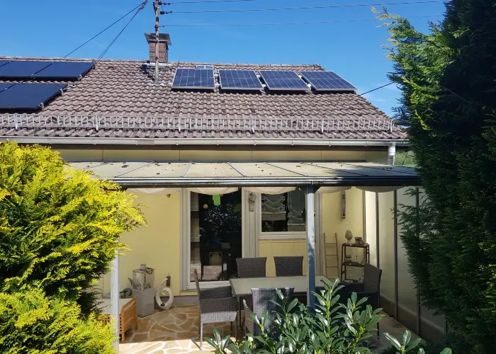 Small house with solar panels, covered patio, outdoor seating, surrounded by green shrubs under clear blue sky
