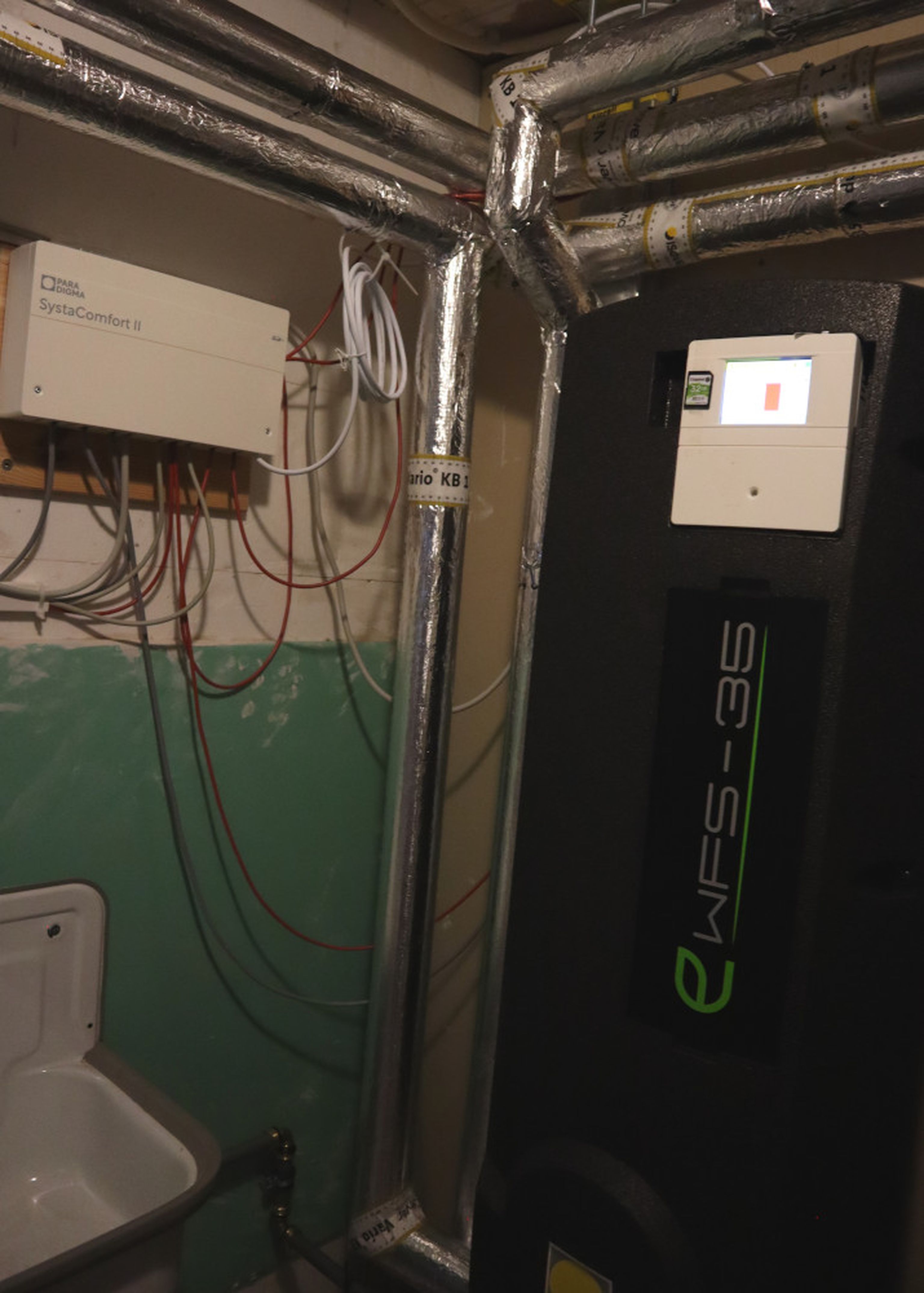 Indoor heating system with insulated pipes, a digital control panel, and an electrical unit. Walls are painted green and white.