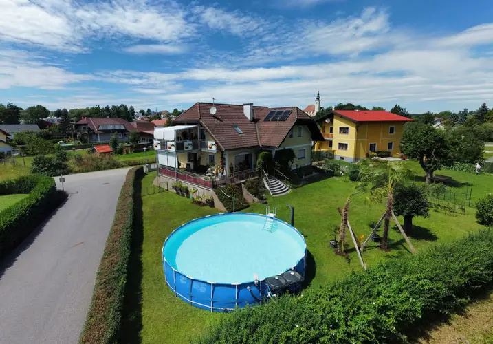 Aerial view of a house with red roof, large backyard, round above-ground pool, surrounded by green lawn and hedges