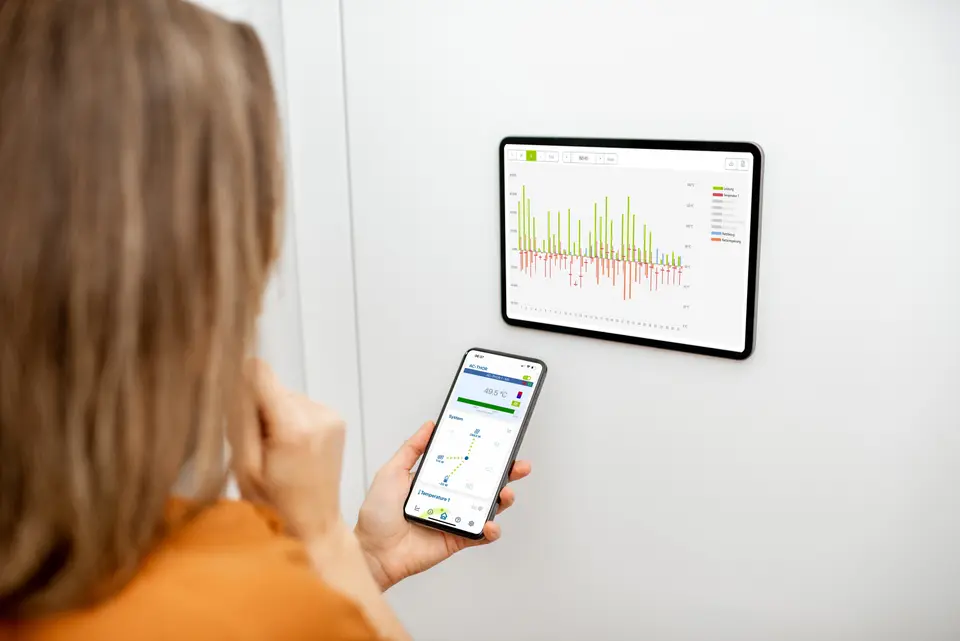 A woman analyzes data charts with green and red bars on a wall-mounted tablet and smartphone.