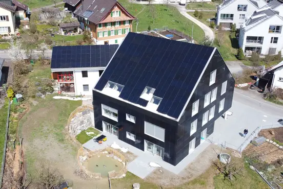 Aerial view of a modern house with solar panels on the roof, surrounded by grass and neighboring houses in a residential area.