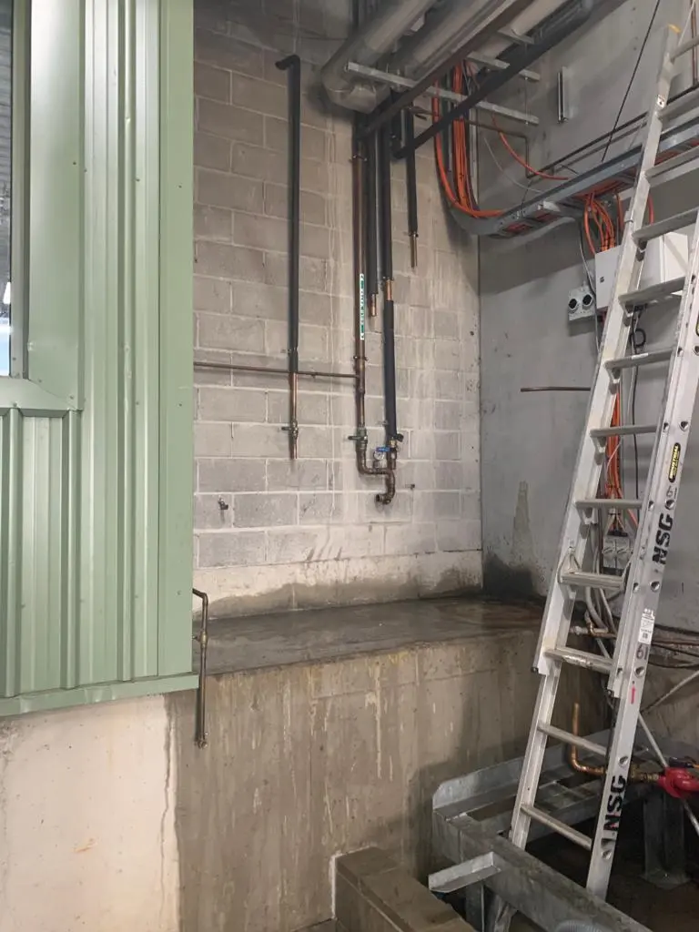 Industrial area with exposed pipes on concrete wall, metal ladder, orange cables, and a green metal panel on the left.
