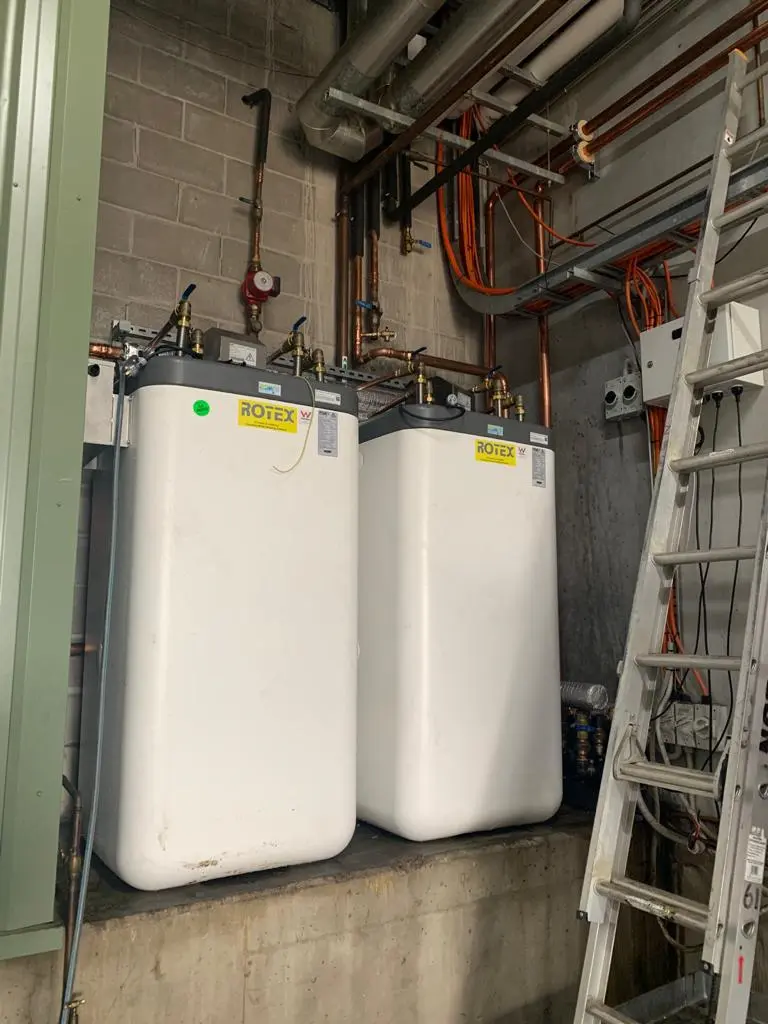 Two large white water heaters with pipes and wiring in a utility room beside a ladder and concrete wall.