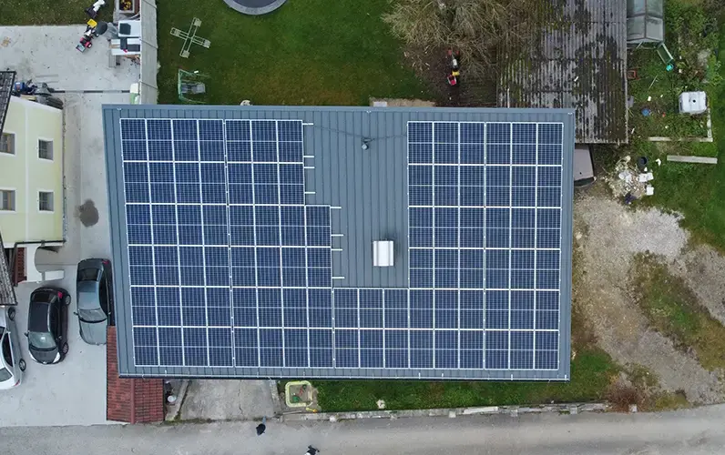 Aerial view of a rooftop covered with solar panels, surrounded by grass, a road, and parked cars.