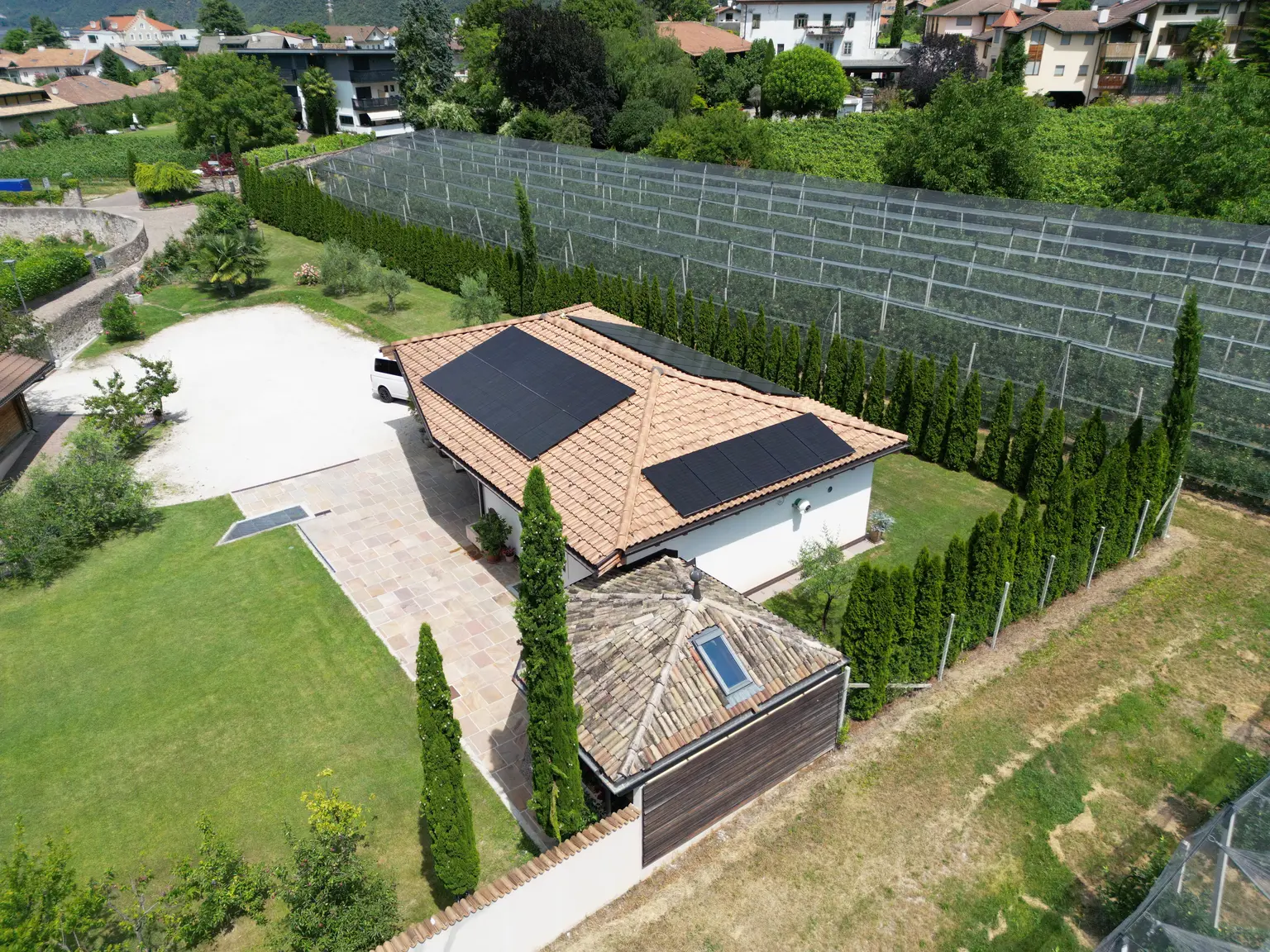 Aerial view of a house with solar panels, surrounded by trees and a large netted area in a residential neighborhood.