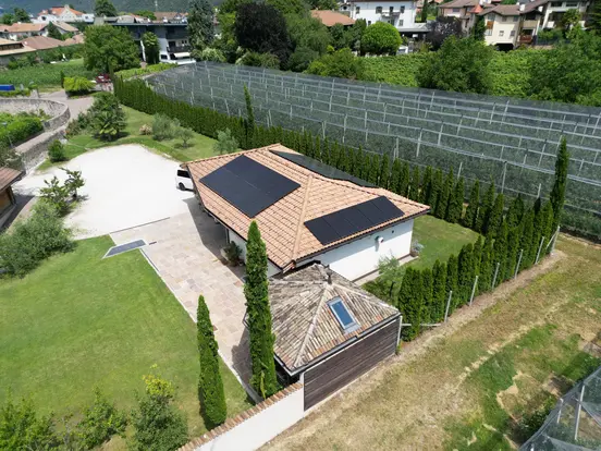 Aerial view of a house with solar panels on the roof, surrounded by green lawn, trees, and a fenced garden area.