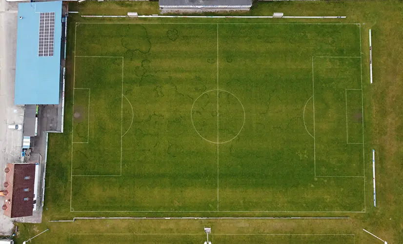 Aerial view of an empty soccer field with goalposts and a blue-roofed building with solar panels on the left