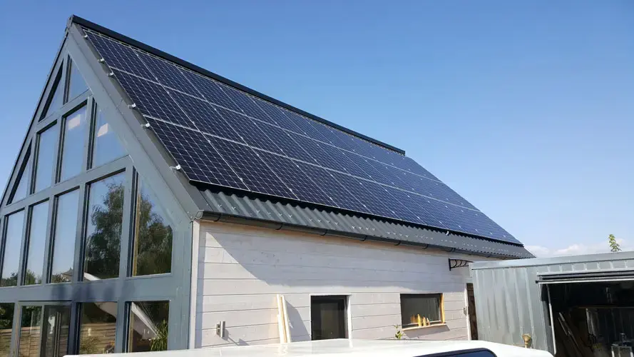 Modern house with large solar panels on roof, glass facade, under clear blue sky.