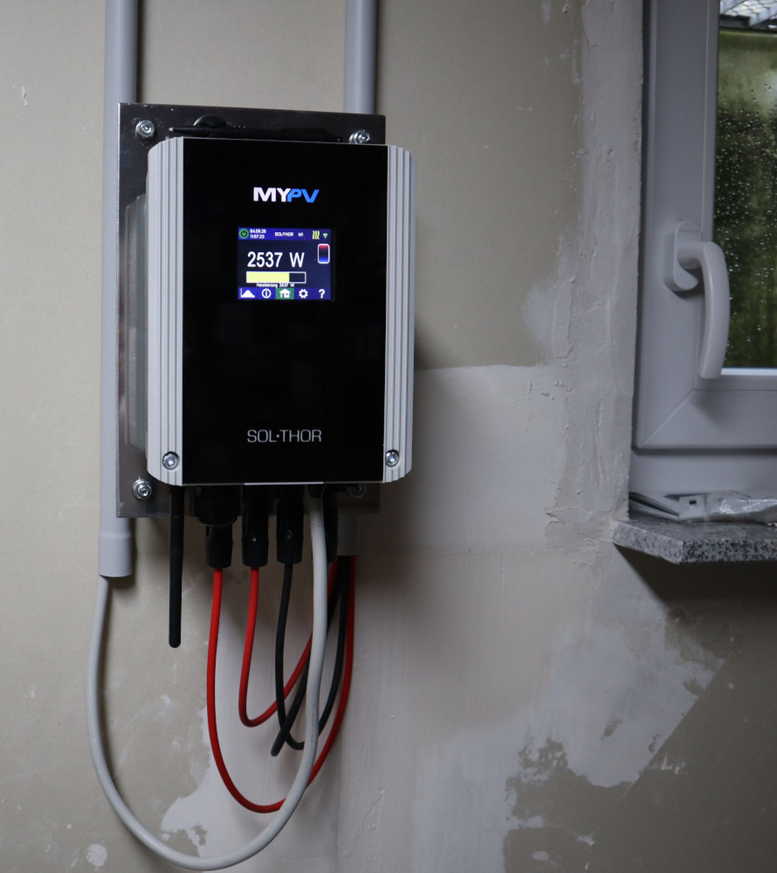 Wall-mounted solar power inverter showing 2537 W output, with wires connected below. A window is partially visible on the right.