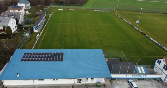 Aerial view of a football field with a blue-roofed building with solar panels, surrounded by houses and greenery