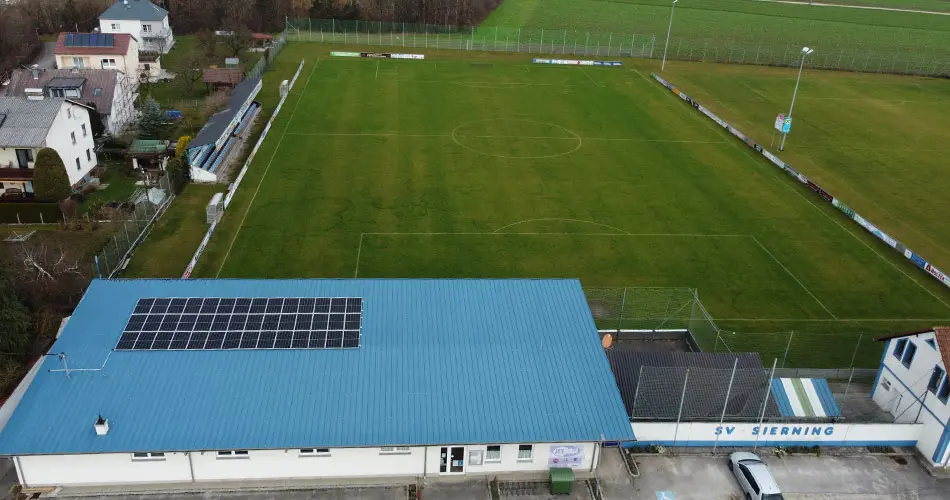 Aerial view of a football field with a blue-roofed building with solar panels, surrounded by houses and greenery