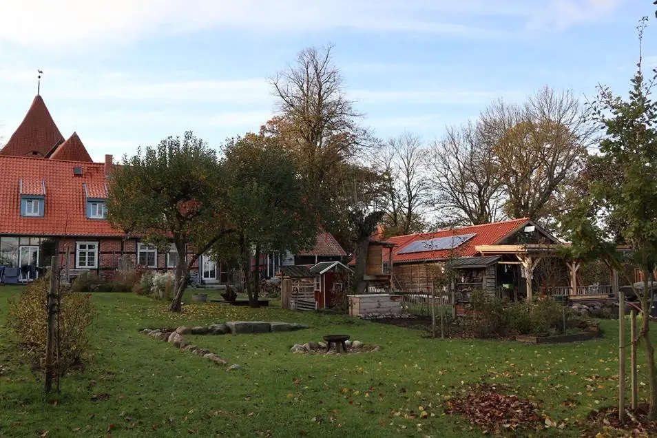 Quaint rural scene with red-roofed house, green lawn, trees, and wooden shed with solar panels under partly cloudy sky