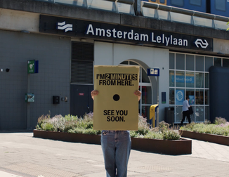 Person holding a sign that reads "I'm 2 minutes from here. See you soon." in front of Amsterdam Lelylaan station.