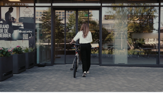 A woman with a bike approaches a glass door entrance to a modern building, with a coffee advertisement visible to the left.
