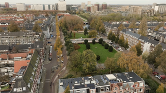 Aerial view of a Amsterdam West with tree-lined streets, red-roofed buildings, grassy park area, and distant high-rise buildings under a clear sky.