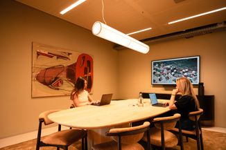 Two women sitting at a conference table with laptops, a large abstract painting on the wall, and a screen displaying a beach scene.