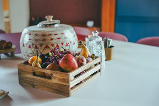 A wooden crate filled with assorted fruits, including apples, pears, and grapes, placed on a table next to a decorative jar and glassware.