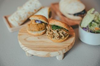 Two small sandwiches on a wooden stand, filled with shredded meat and greens, with wraps and salad in the background.