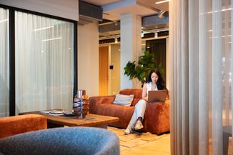 A woman sits on a brown sofa, using a laptop in a modern, cozy room with a coffee table, plant, and large windows.
