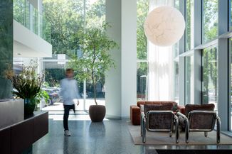 Modern lobby with large windows, greenery, brown seating, a round hanging light, and a blurred person walking, with trees visible outside.
