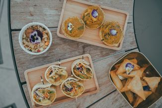 Assorted vegetarian dishes on wooden trays, including wraps, pastries, and a bowl of grains, each garnished with edible flowers.