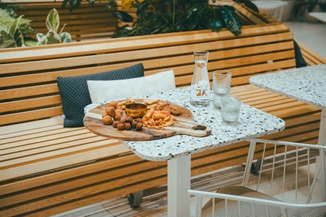 A terrazzo table with a wooden platter of snacks, water carafe, and glasses, set beside a wooden bench with cushions in a cozy outdoor setting.