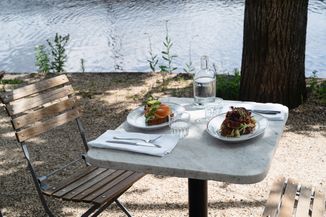 Outdoor table by a lake with two plates of food, cutlery on napkins, and glasses next to a bottle of water on a sunny day.