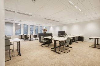 Modern office with several desks and chairs, large windows, neutral colors, and a bouquet of flowers on a cabinet under soft lighting.