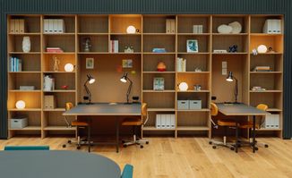 Modern office space with wooden shelves, lamps, and two desks, surrounded by plants. A glass-walled upper floor is visible above.