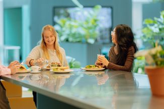Three women enjoy lunch and drinks at a modern restaurant table, surrounded by greenery.