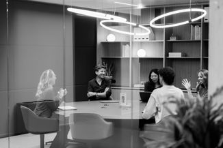 Black and white image of five people having a meeting in a modern office, seated around a round table, discussing and smiling.