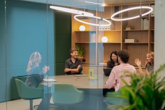 A group of people having a meeting in a modern office with bookshelves, indoor plants, and circular ceiling lights.