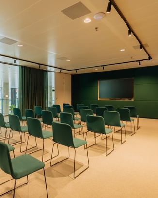 Modern conference room with rows of green chairs facing a large screen, natural light from tall windows, and a beige carpet.